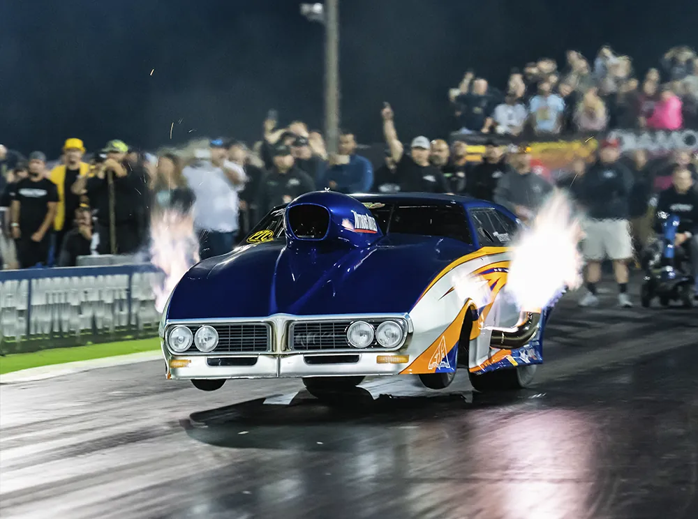 Pro Mod azul largando à noite com fogo nos escapamentos e público atrás da grade